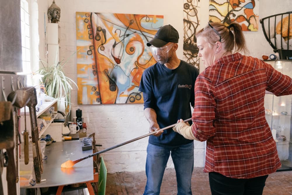 a woman being shown how to do glassblowing in a workshop on a glassblowing experience