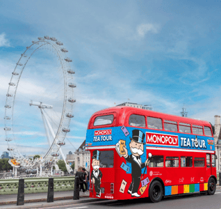a red routemaster bus with colourful monopoly tea tour signs on it with the london eye in the background