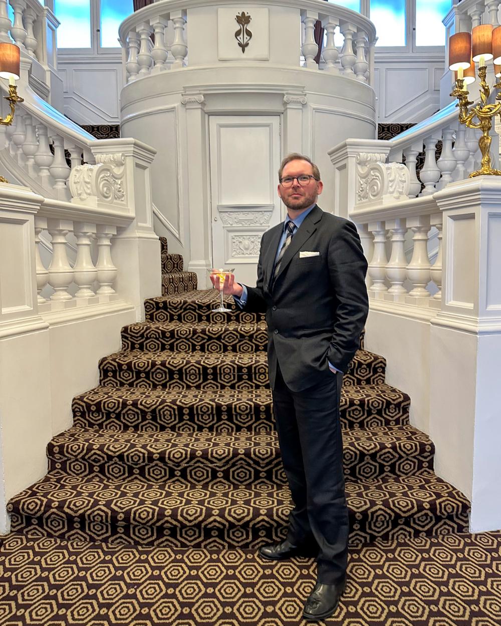 man wearing smart suit holding a martini in front of a grand staircase as part of james bond tour