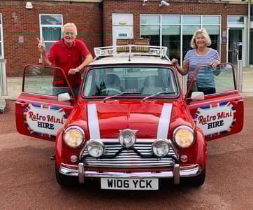 a man and a woman standing either side of a classic red mini cooper on a self drive road trip