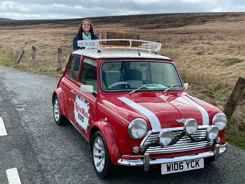 a woman standing behind a classic red mini cooper on a self drive road trip