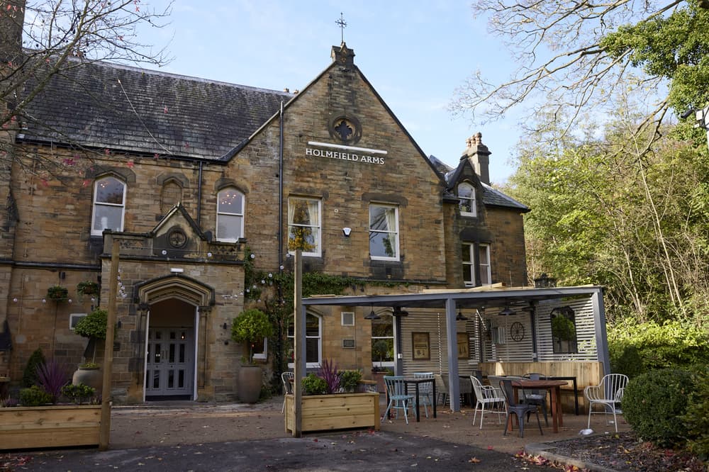 an outside shot of the holmfield arms, a rustic brick building with outdoor seating area with trees surrounding the building