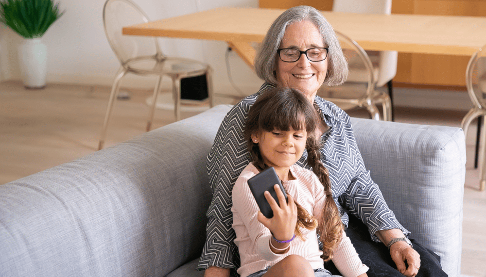 a grandma sitting next to a child on a sofa looking at a smartphone on mother's day