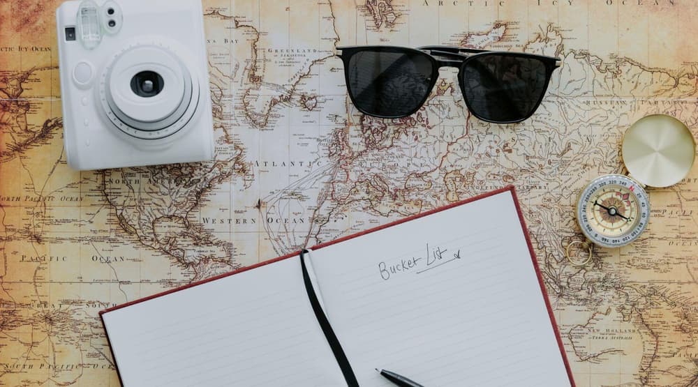 A map with a compass black sunglasses a white camera and a notebook open on top of it with bucket list written on the page