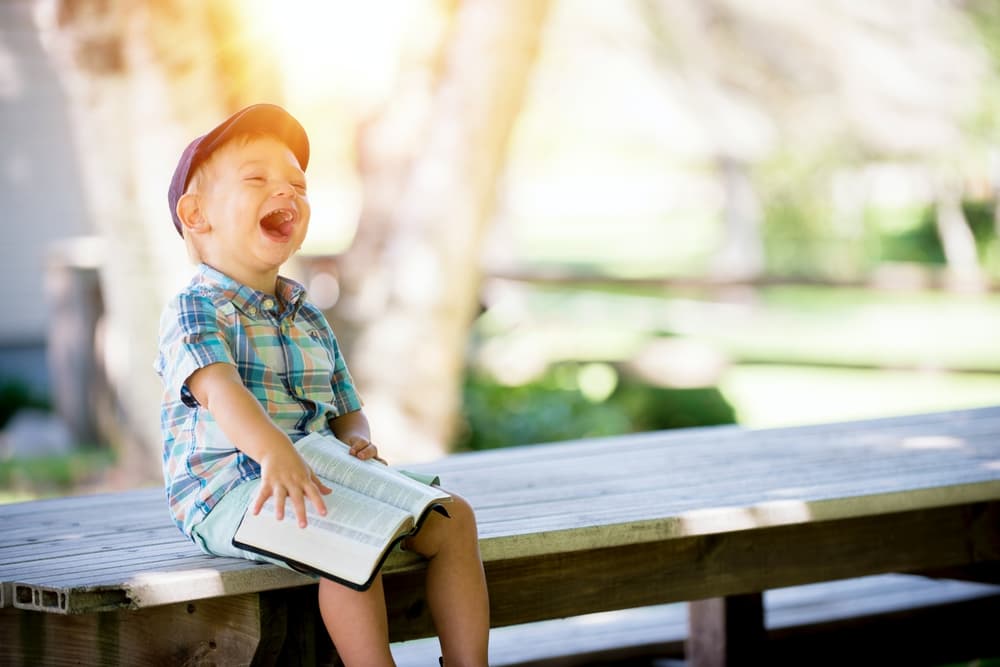 small boy sitting on a bench holding a book and laughing in the sunshine