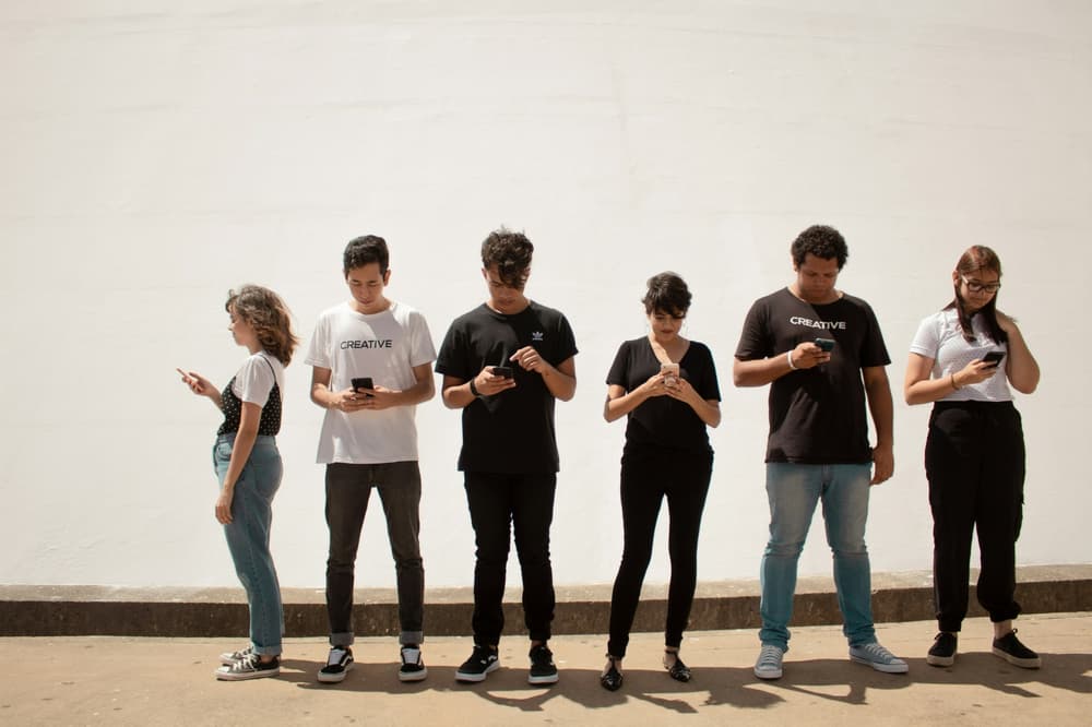 six people in a row against a white wall all looking at their phones