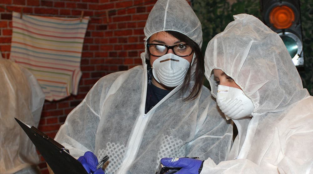 two people dressed in personal protective clothing and wearing face masks taking part in a csi investigation experience day