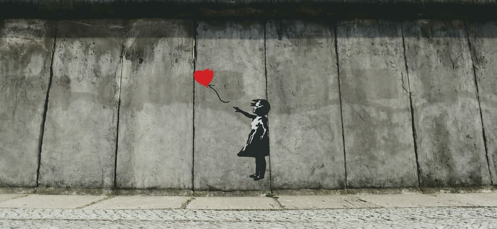 banksy street art on a concrete wall of a young girl letting go of a red heart balloon