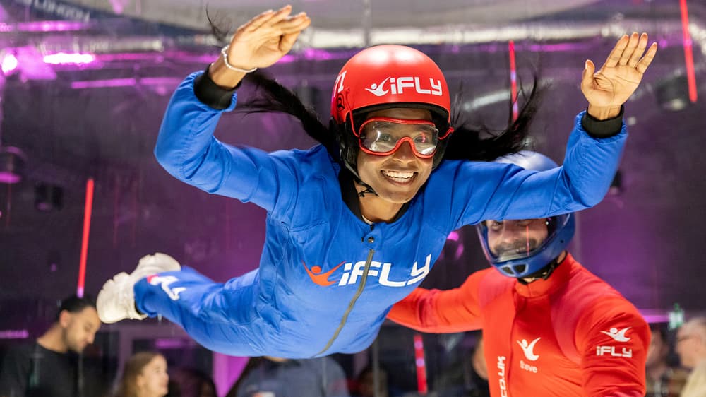 a woman wearing a blue flight suit, goggles and helmet being helped by an instructor in a red suit on an indoor skydiving experience
