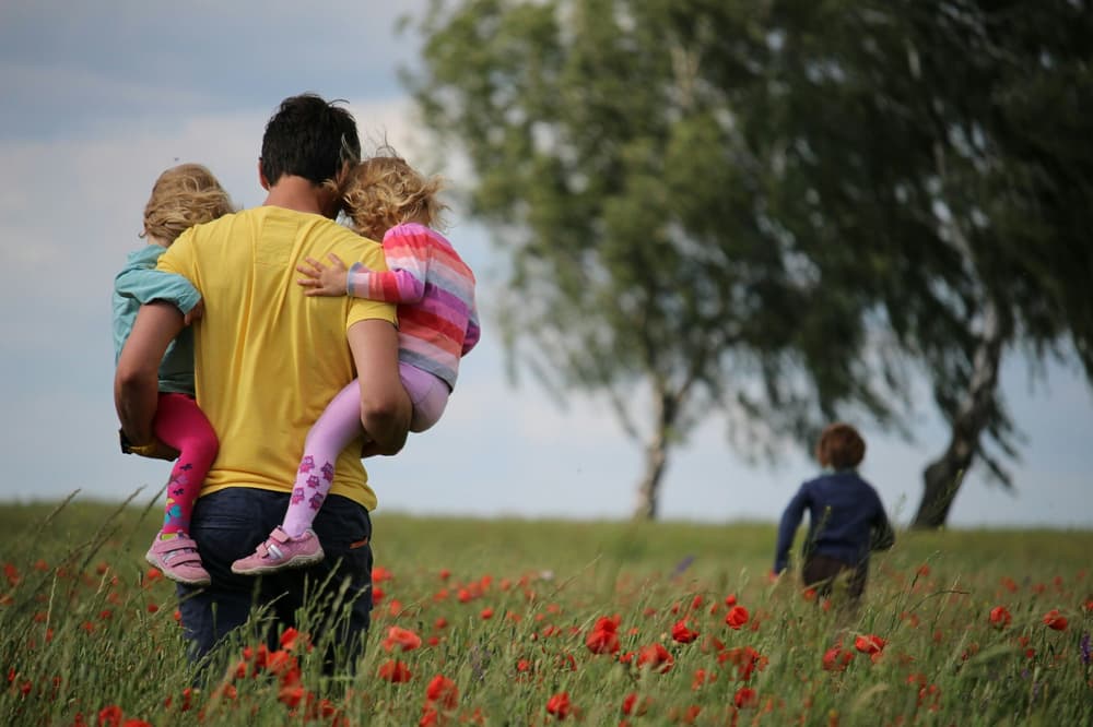 man in a yellow t-shirt carrying 2 children through a field of poppies with another child running ahead