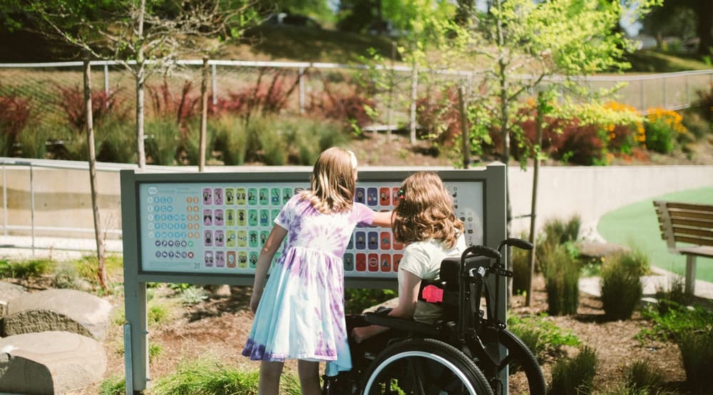 Two young girls one in a wheelchair in a park looking at a sign