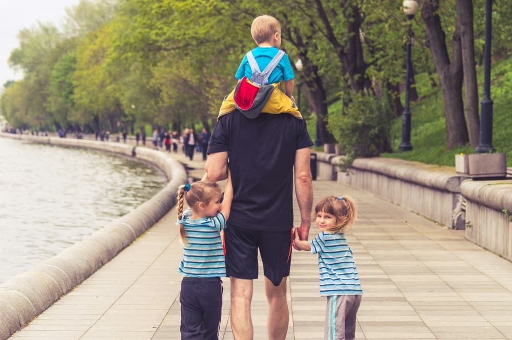 man with hs back to the camera holding hands with two small children and carrying one on his shoulders in public park