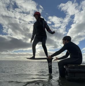 woman standing on a pump foil board with instructor sitting on the side of the water