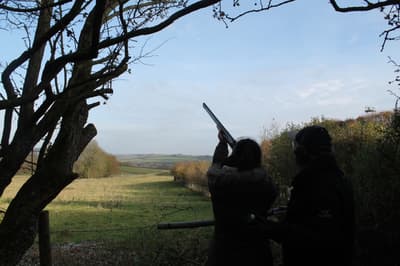 a person aiming a gun into the sky looked on by an instructor taking part in a clay pigeon shooting experience