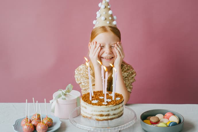 young girl wearing a party hat at a table with a birthday cake and lit candles