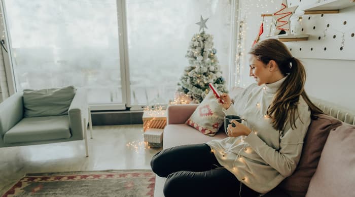 Woman sitting on sofa holding mug and looking at phone with christmas tree in corner of room