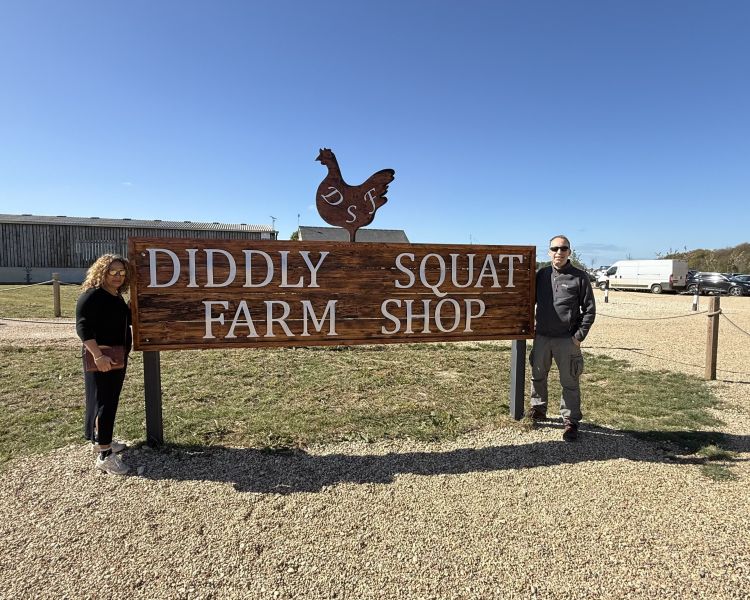 two people next to the diddly squat farm shop sign on clarkson's farm