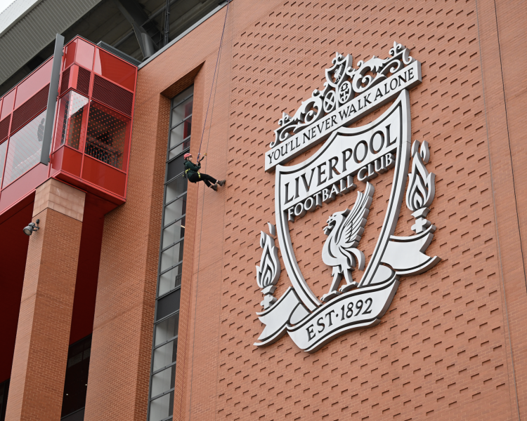 view of person abseiling down the side of anfield stadium past the liverpool fc crest
