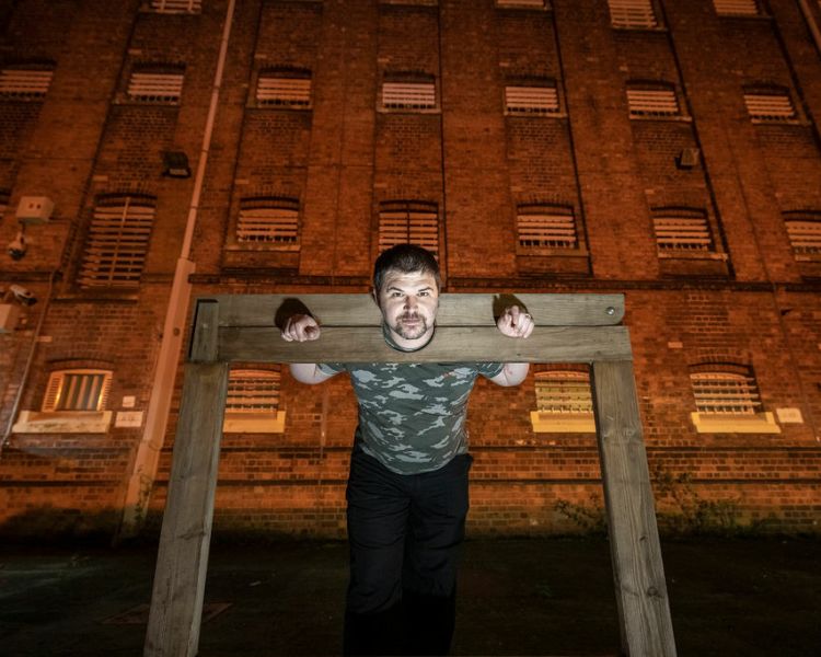 a man with his head and hands in wooden stocks in front of a prison at night as part of a ghost tour at a haunted prison experience day
