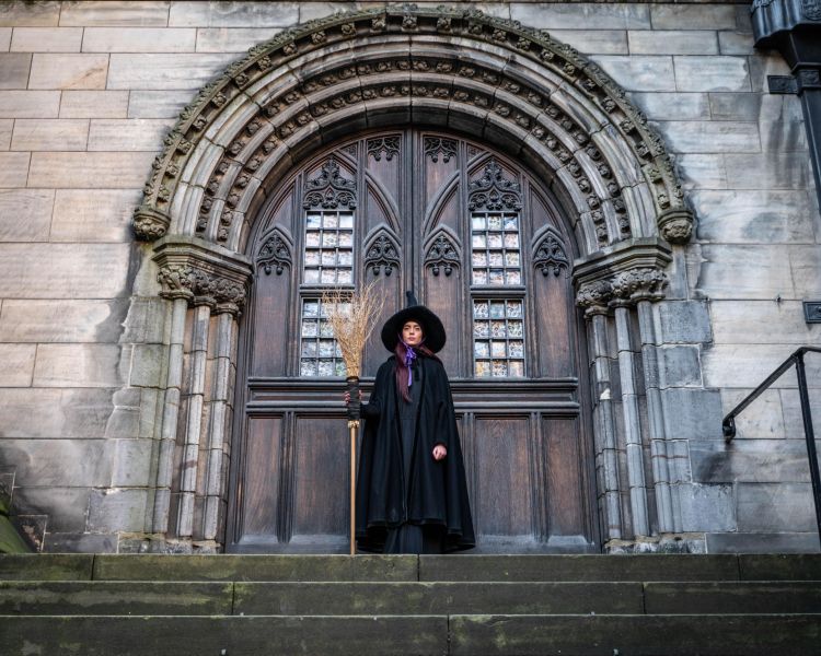 guide dressed as witch in front of big wooden doors on a witches walking tour in edinburgh