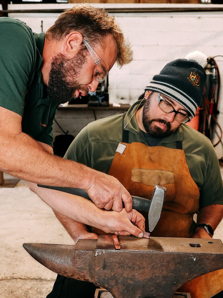 man being shown how to hammer and shape hot metal by instructor on silversmithing course