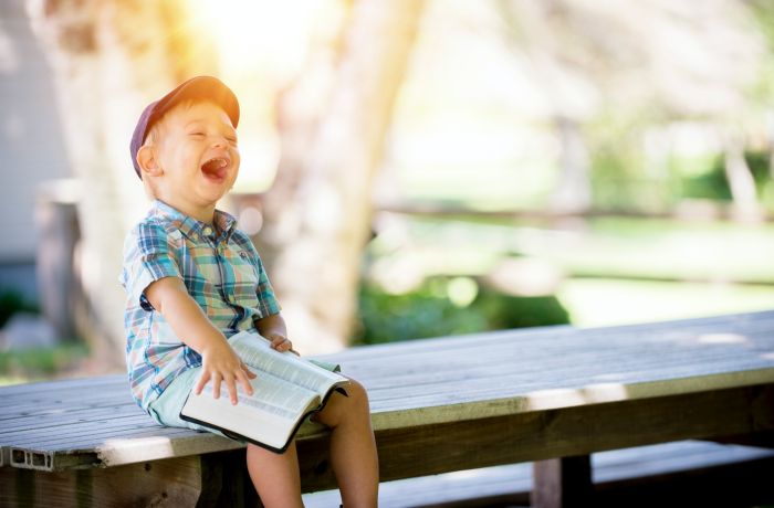 small boy sitting on a bench holding a book and laughing in the sunshine