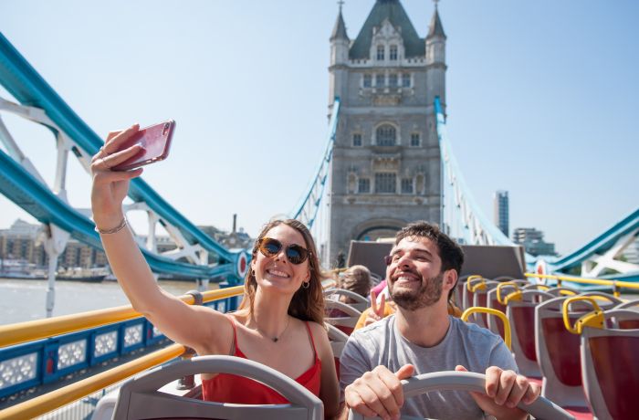 couple on top deck of the original london bus tour taking selfie going over tower bridge