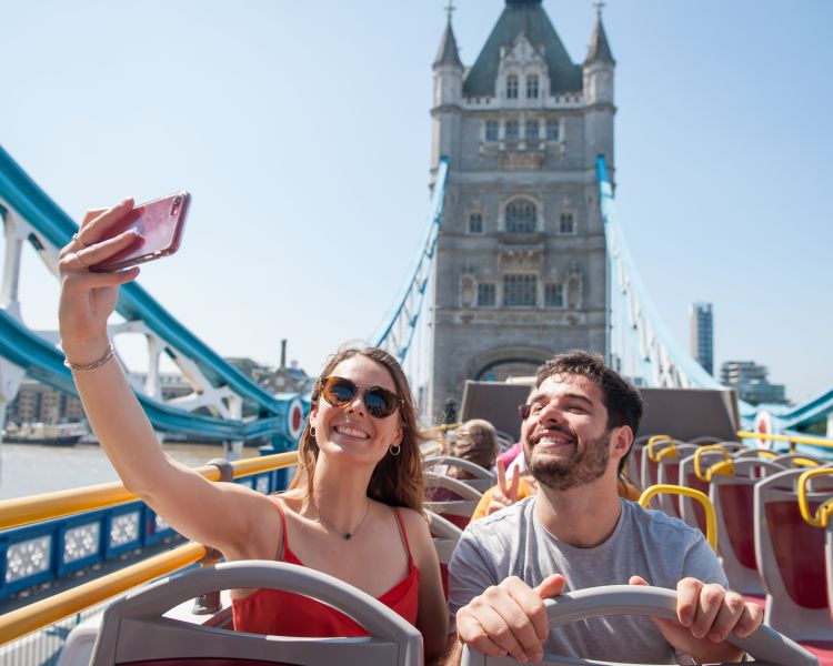 couple on top deck of the original london bus tour taking selfie going over tower bridge