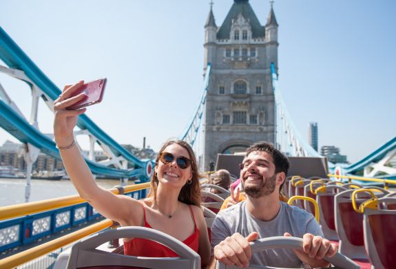 couple on top deck of the original london bus tour taking selfie going over tower bridge