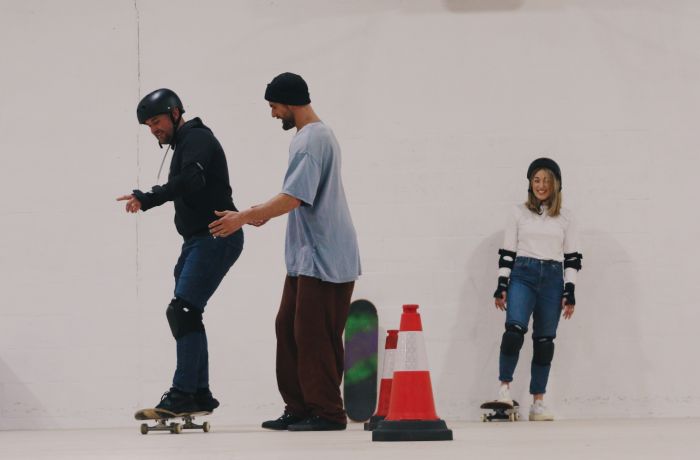 man being helped by an instructor with woman looking on at a skateboard lesson for two