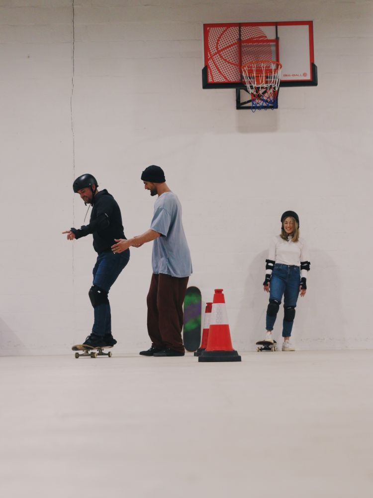 man being helped by an instructor with woman looking on at a skateboard lesson for two