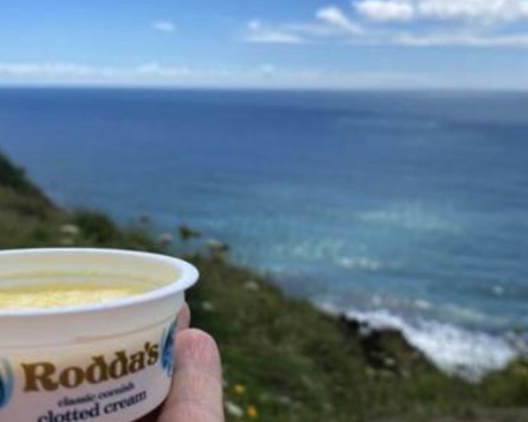 hand holding a pot of rodda's clotted cream overlooking the cornish coastline