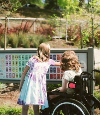 Two young girls one in a wheelchair in a park looking at a sign