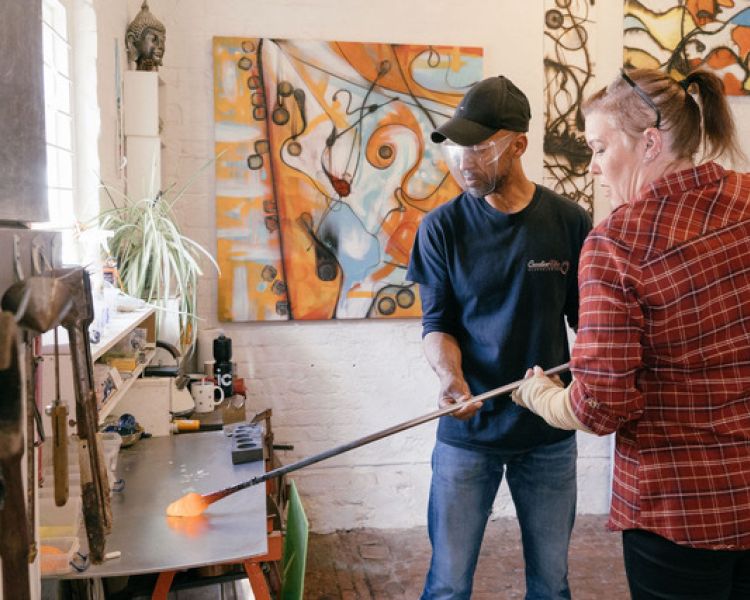 a woman being shown how to do glassblowing in a workshop on a glassblowing experience
