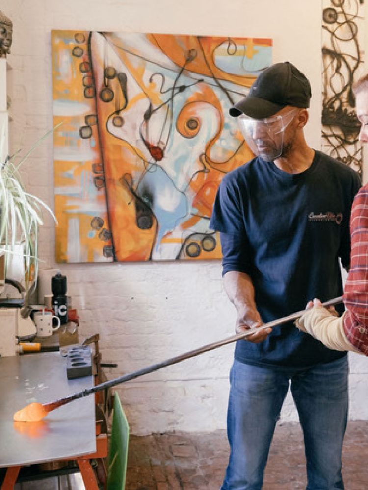 a woman being shown how to do glassblowing in a workshop on a glassblowing experience