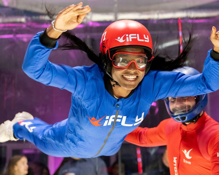 a woman wearing a blue flight suit, goggles and helmet being helped by an instructor in a red suit on an indoor skydiving experience