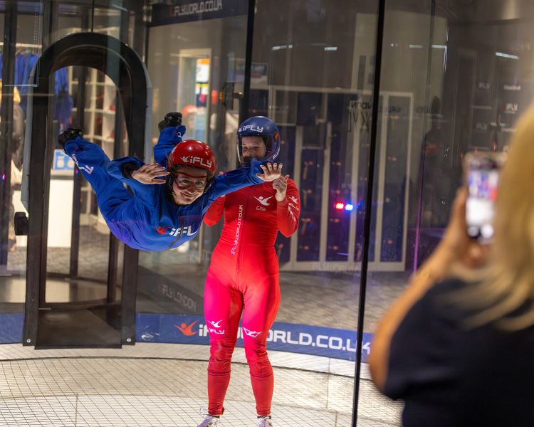 a woman wearing a blue flight suit, goggles and helmet being helped by an instructor in a red suit on an indoor skydiving experience