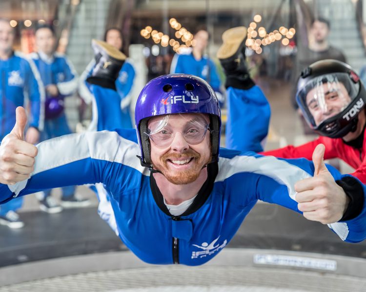 a man wearing a blue flight suit, goggles and helmet giving the thumbs up on an indoor skydiving experience