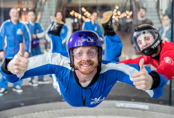 a man wearing a blue flight suit, goggles and helmet giving the thumbs up on an indoor skydiving experience