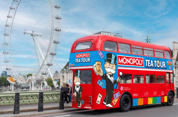 a red routemaster bus with colourful monopoly tea tour signs on it with the london eye in the background