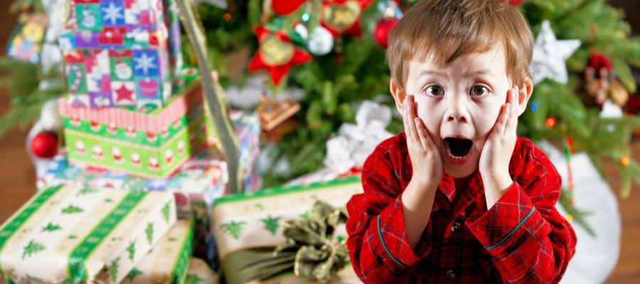 Young boy looking surprised as he stands by a christmas tree with presents underneath