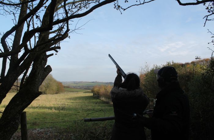 a person aiming a gun into the sky looked on by an instructor taking part in a clay pigeon shooting experience