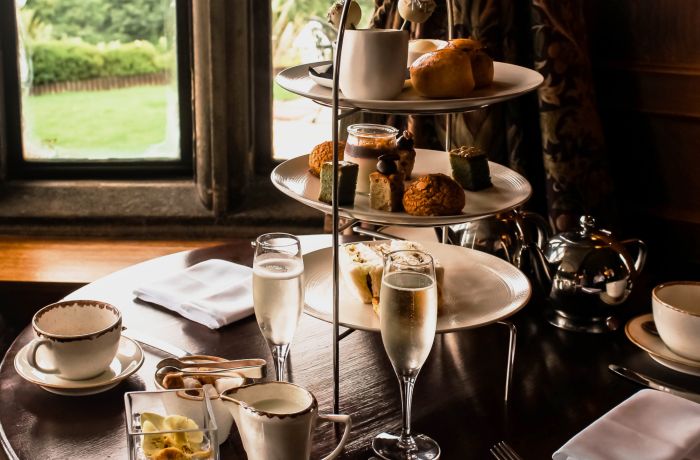 an afternoon tea set up in a restaurant on a round wooden table next to a window with champagne and cake stand full of cakes and sandwiches