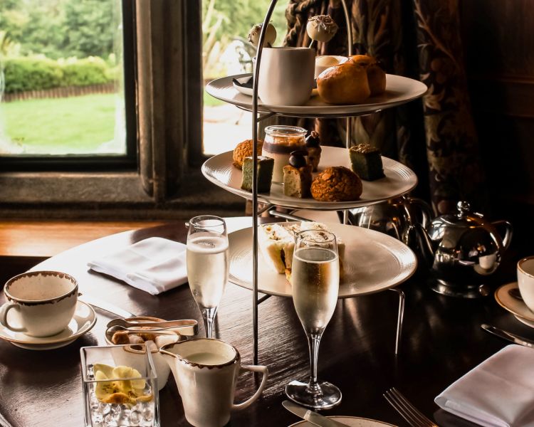 an afternoon tea set up in a restaurant on a round wooden table next to a window with champagne and cake stand full of cakes and sandwiches