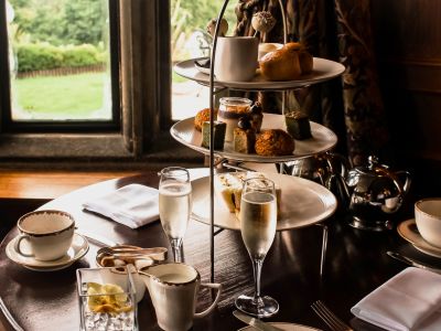 an afternoon tea set up in a restaurant on a round wooden table next to a window with champagne and cake stand full of cakes and sandwiches