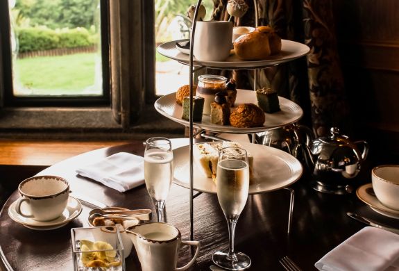 an afternoon tea set up in a restaurant on a round wooden table next to a window with champagne and cake stand full of cakes and sandwiches