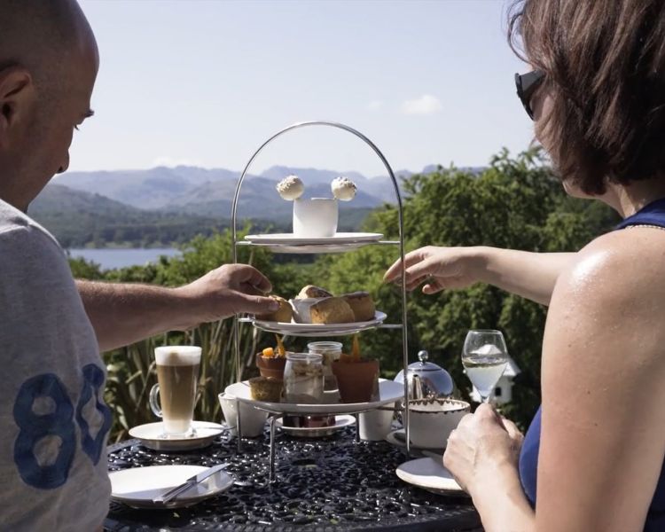 a couple enjoying an afternoon tea on a terrace overlooking a lake in the Lake District on a sunny day