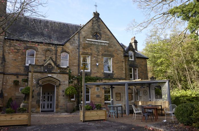 an outside shot of the holmfield arms, a rustic brick building with outdoor seating area with trees surrounding the building