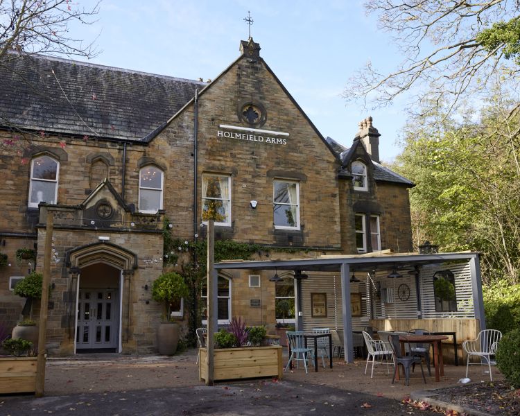 an outside shot of the holmfield arms, a rustic brick building with outdoor seating area with trees surrounding the building