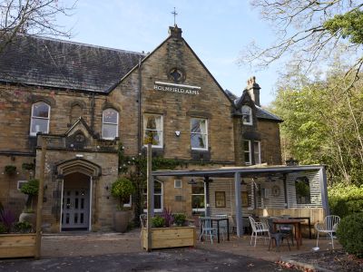 an outside shot of the holmfield arms, a rustic brick building with outdoor seating area with trees surrounding the building